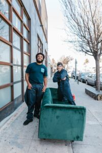 Two movers in work attire relocate a green couch on a city sidewalk.